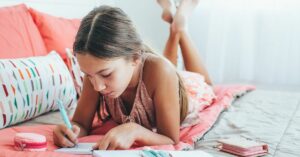 a young girl lies on a bed and writes in her journal