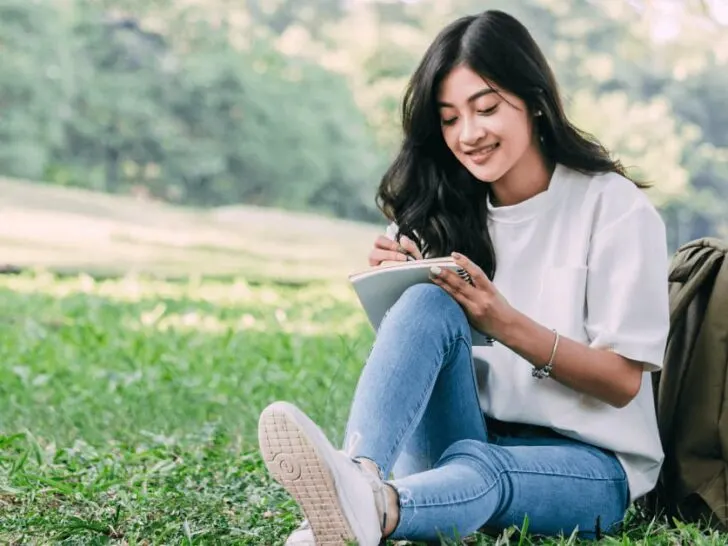 a teen girl sits on grass and journals in a notebook