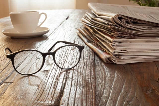 a stack of newspapers, a white cup, and a pair of glasses